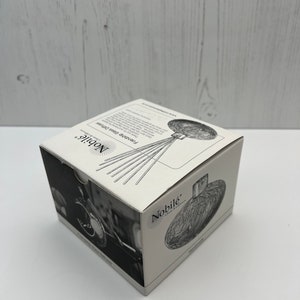 May include: A white box with a black and white image of a glass bottle stopper with a floral design. The box has the text "Nobil&eacute;" on it. The bottle stopper is a decorative item for wine bottles.