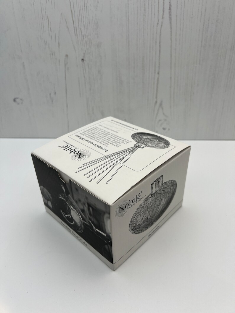 May include: A white box with a black and white image of a glass bottle stopper with a floral design. The box has the text "Nobil&eacute;" on it. The bottle stopper is a decorative item for wine bottles.