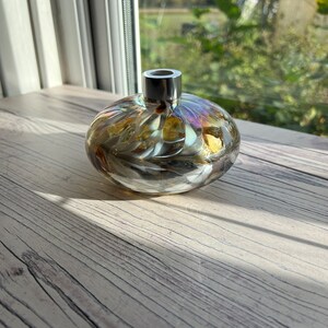 May include: A round glass bottle with a silver top. The bottle has a swirling pattern of gold, white, and iridescent colors. The bottle is sitting on a light wood surface.