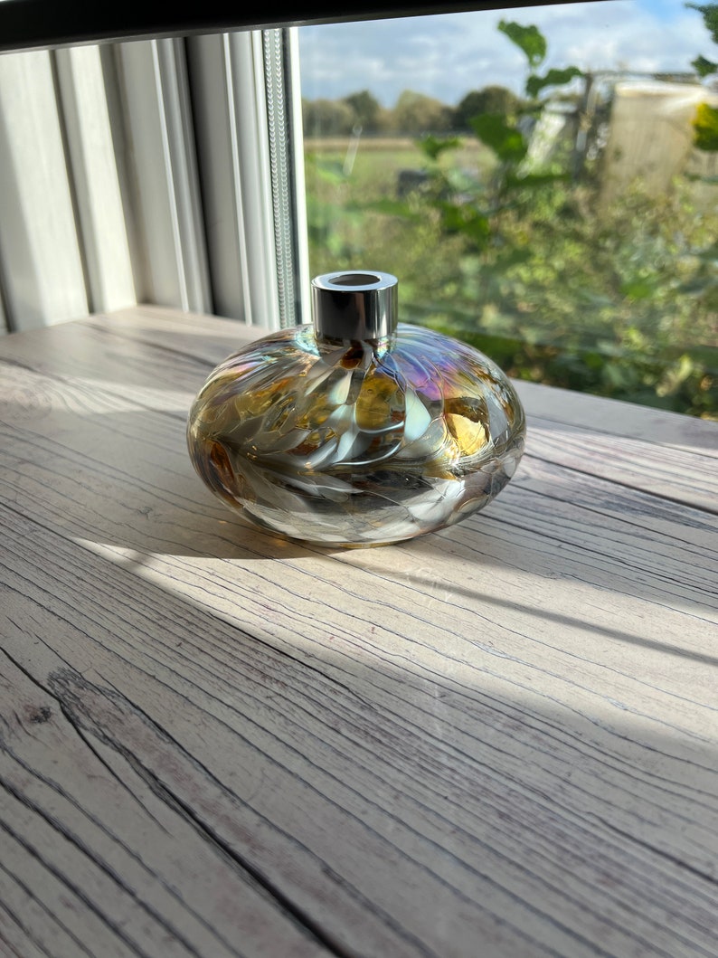 May include: A round glass bottle with a silver top. The bottle has a swirling pattern of gold, white, and iridescent colors. The bottle is sitting on a light wood surface.