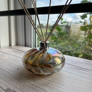 May include: A diffuser bottle with a swirl pattern in shades of gold, white, and brown. The bottle has a silver cap and is filled with reeds.