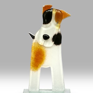 Puede incluir: Una figurita de vidrio de un perro blanco, marrón y negro con un patrón moteado. El perro está de pie sobre una base de vidrio transparente.