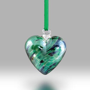 Può includere: Un ornamento a forma di cuore in vetro verde con vortici di nero e oro. L'ornamento è appeso a un nastro verde.
