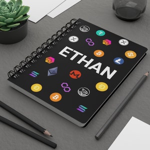 Puede incluir: Una libreta negra con encuadernación en espiral y el nombre "ETHAN" escrito en letras blancas. La libreta está decorada con varios logotipos de criptomonedas de colores, incluyendo Bitcoin, Ethereum, Litecoin y otros.