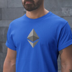 Puede incluir: Una camiseta azul real con un logotipo gris de criptomonedas Ethereum impreso en la parte delantera.