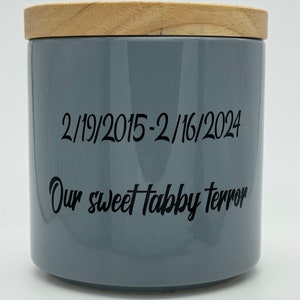 May include: A gray ceramic jar with a wooden lid. The jar has black text that reads "2/19/2015-2/16/2024 Our sweet tabby terror".