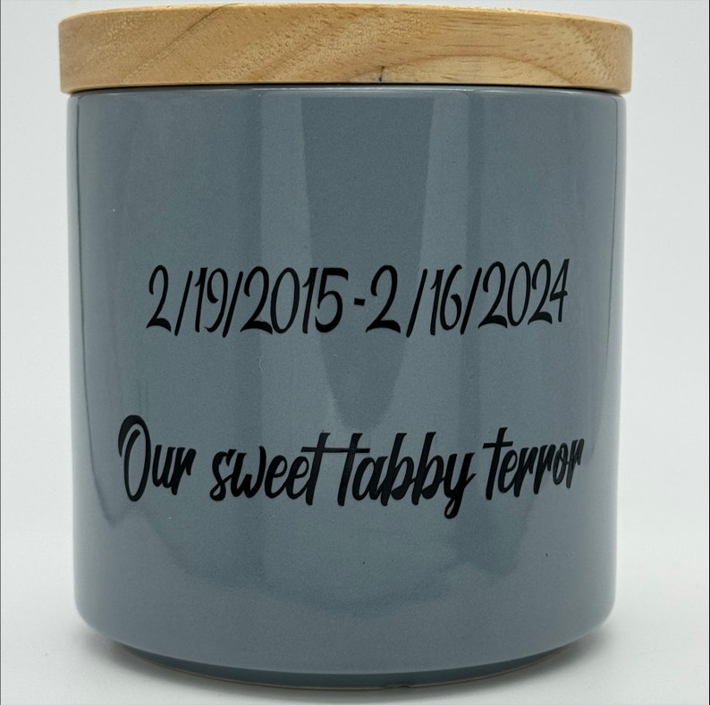 May include: A gray ceramic jar with a wooden lid. The jar has black text that reads "2/19/2015-2/16/2024 Our sweet tabby terror".