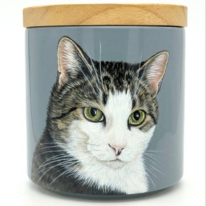 May include: A ceramic jar with a wooden lid features a hand-painted portrait of a tabby cat with white markings. The cat has green eyes and a serious expression. The jar is painted on a gray background.