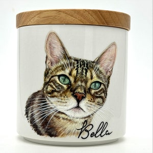 May include: A white ceramic jar with a wooden lid, featuring a hand-painted portrait of a tabby cat with green eyes. The cat's name, "Bella", is written in cursive below the image.