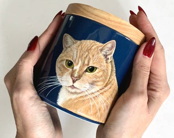 May include: A ceramic mug with a wooden lid features a hand-painted portrait of a ginger cat with green eyes. The cat is looking directly at the viewer.