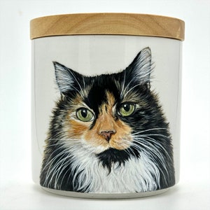 May include: A white ceramic jar with a wooden lid features a hand-painted portrait of a calico cat with green eyes. The cat's fur is black, white, and orange.