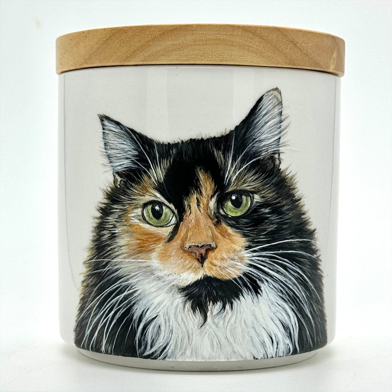 May include: A white ceramic jar with a wooden lid features a hand-painted portrait of a calico cat with green eyes. The cat's fur is black, white, and orange.