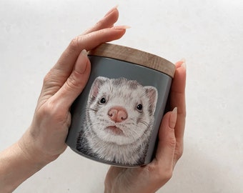 May include: A gray ceramic jar with a wooden lid features a hand-painted illustration of a ferret. The ferret has white fur with black markings around its eyes and nose. The ferret is looking directly at the viewer.