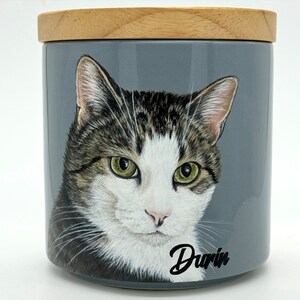 May include: A gray ceramic jar with a wooden lid, featuring a hand-painted portrait of a tabby cat with green eyes. The cat's name, "Darin", is written in black cursive script on the jar.