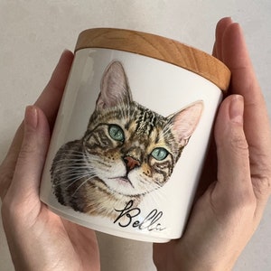 May include: A white ceramic jar with a wooden lid, featuring a hand-painted portrait of a tabby cat with green eyes. The cat's name, "Bella", is written in cursive below the image.