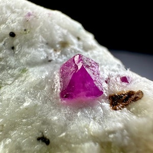 Può includere: Un cristallo di rubino rosa vibrante, con una forma geometrica appuntita, si trova su una roccia bianca e soffice. Il rubino è il punto focale dell'immagine, con la roccia che fornisce uno sfondo contrastante.