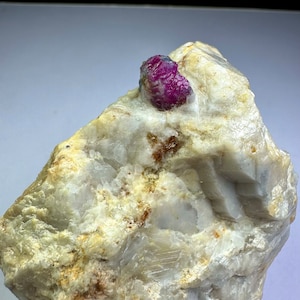 Könnte beinhalten: Ein leuchtend rosa Rubin-Edelstein, eingebettet in einen rauen weißen Stein. Der Rubin hat eine tiefrosa Farbe und eine glatte, polierte Oberfläche. Der Stein ist hellgrau und hat eine raue, strukturierte Oberfläche.