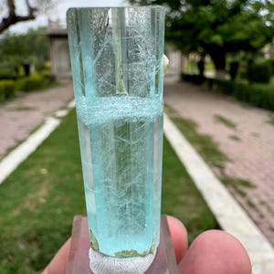 Pu&ograve; includere: Cristallo di acquamarina blu chiaro, traslucido, montato su una base rettangolare trasparente. Il cristallo presenta venature interne e una consistenza leggermente ruvida alla base. Lo sfondo mostra una scena esterna sfocata con vegetazione.