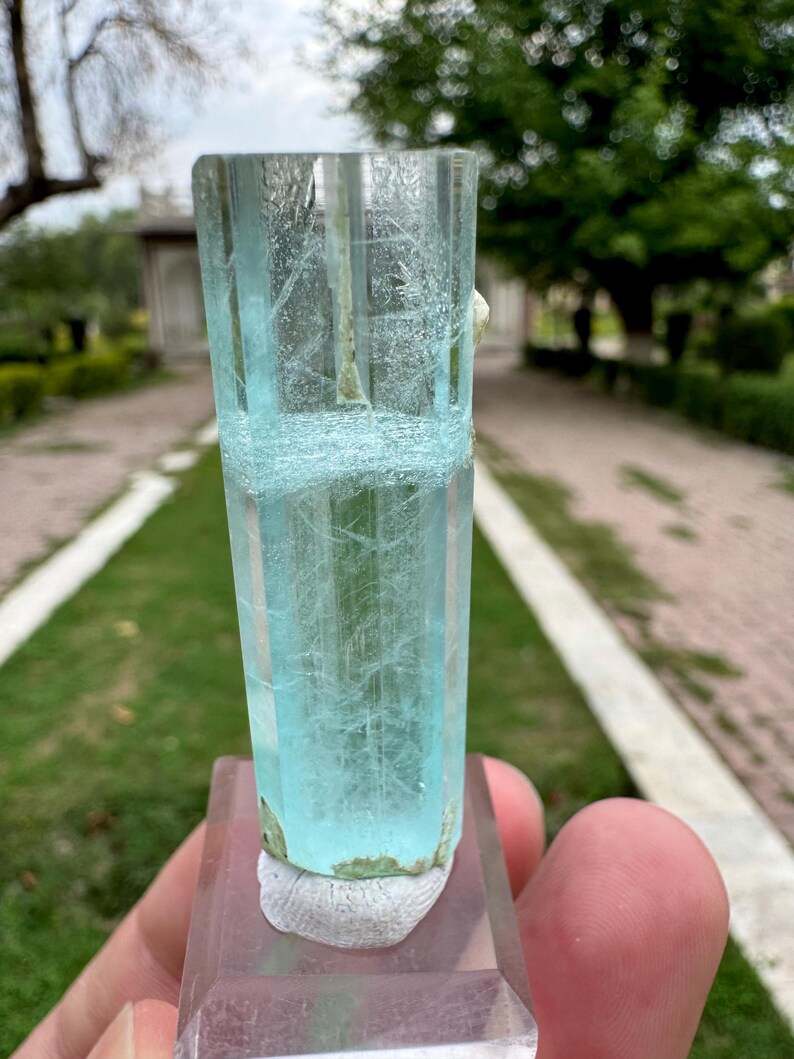 Pu&ograve; includere: Cristallo di acquamarina blu chiaro, traslucido, montato su una base rettangolare trasparente. Il cristallo presenta venature interne e una consistenza leggermente ruvida alla base. Lo sfondo mostra una scena esterna sfocata con vegetazione.