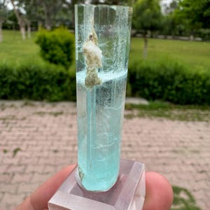 Pu&ograve; includere: Cristallo di acquamarina azzurra, montato su una base rettangolare trasparente. Il cristallo &egrave; un prisma traslucido e allungato con una consistenza naturale ruvida. Un'inclusione bianca e beige &egrave; visibile all'interno del cristallo. Lo sfondo &egrave; una scena esterna sfocata.