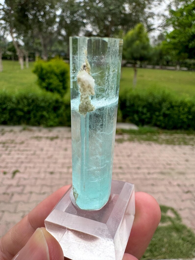 Pu&ograve; includere: Cristallo di acquamarina azzurra, montato su una base rettangolare trasparente. Il cristallo &egrave; un prisma traslucido e allungato con una consistenza naturale ruvida. Un'inclusione bianca e beige &egrave; visibile all'interno del cristallo. Lo sfondo &egrave; una scena esterna sfocata.