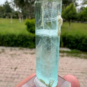 Pu&ograve; includere: Cristallo rettangolare traslucido di acquamarina, di colore azzurro chiaro. Il cristallo ha una superficie ruvida e strutturata, montato su una base quadrata trasparente. Il cristallo &egrave; tenuto in mano, con uno sfondo verde sfocato.