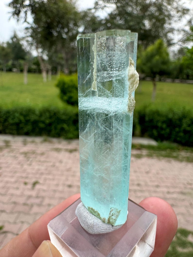 Pu&ograve; includere: Cristallo rettangolare traslucido di acquamarina, di colore azzurro chiaro. Il cristallo ha una superficie ruvida e strutturata, montato su una base quadrata trasparente. Il cristallo &egrave; tenuto in mano, con uno sfondo verde sfocato.