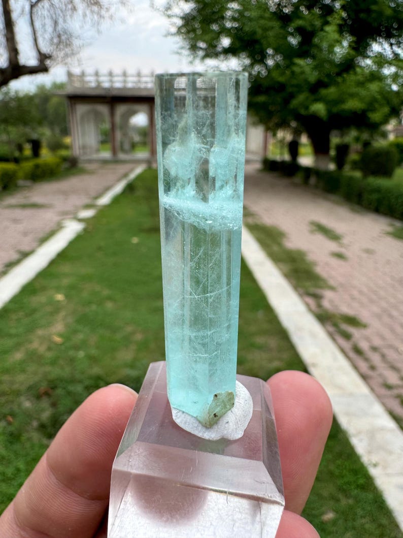 Pu&ograve; includere: Cristallo rettangolare trasparente con una tonalit&agrave; azzurra, montato su una base trasparente. Il cristallo presenta fratture e inclusioni interne. Un piccolo minerale verdognolo &egrave; attaccato alla base. Sfondo di un giardino.