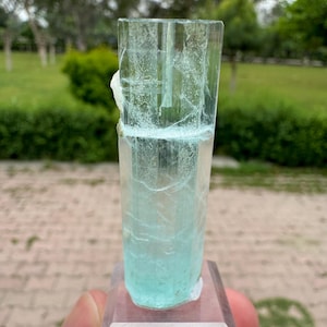 Pu&ograve; includere: Un cristallo trasparente e rettangolare con una tonalit&agrave; azzurra, montato su una base trasparente. Il cristallo presenta fratture interne e un'area ruvida e bianca su un lato. Lo sfondo &egrave; una scena esterna sfocata con fogliame verde.