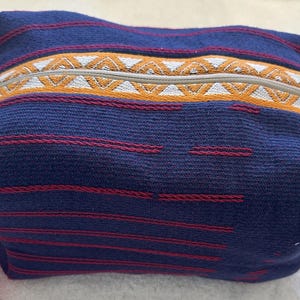 May include: A rectangular fabric pouch in deep blue with thin horizontal red stripes. A central band features a geometric pattern in orange and white, with a light-colored zipper running along the top. The pouch has a soft, woven texture.