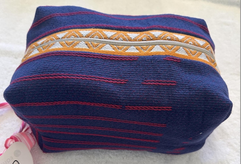 May include: A rectangular fabric pouch in deep blue with thin horizontal red stripes. A central band features a geometric pattern in orange and white, with a light-colored zipper running along the top. The pouch has a soft, woven texture.