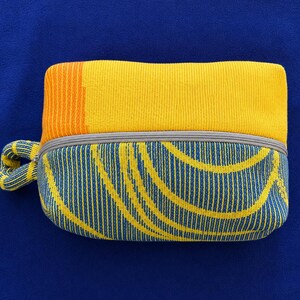 May include: A rectangular zippered pouch featuring a knit texture. The top section is solid yellow with an orange vertical stripe on the left. The bottom section displays a blue and yellow curved line pattern. A small matching knit loop is attached.