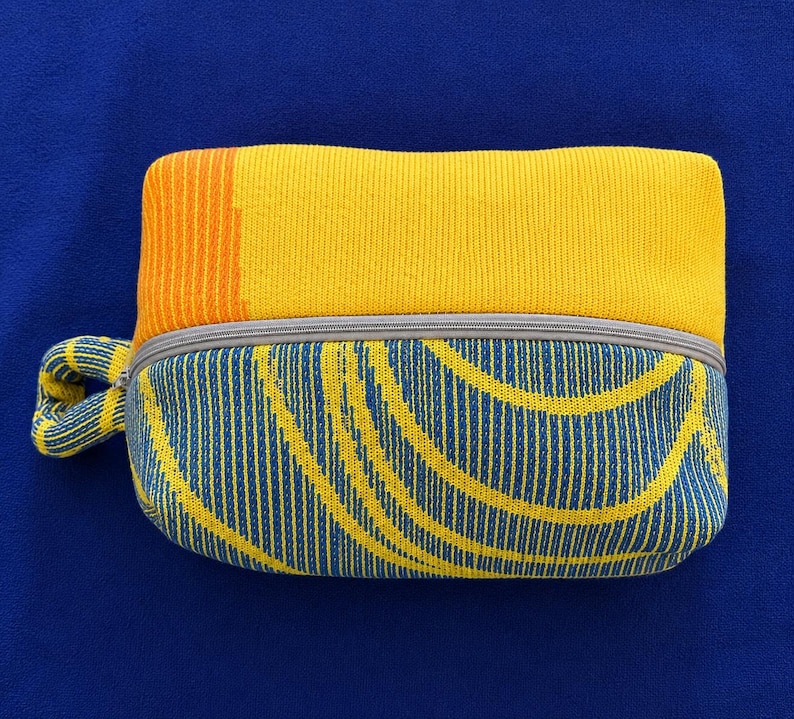 May include: A rectangular zippered pouch featuring a knit texture. The top section is solid yellow with an orange vertical stripe on the left. The bottom section displays a blue and yellow curved line pattern. A small matching knit loop is attached.