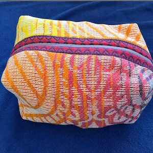 May include: A rectangular zippered pouch featuring a vibrant woven fabric with a multi-colored pattern of orange, yellow, and pink stripes. The top edge is accented with a dark purple trim featuring a repeating geometric pattern along the zipper.