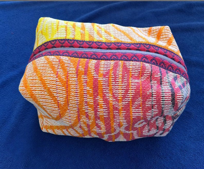 May include: A rectangular zippered pouch featuring a vibrant woven fabric with a multi-colored pattern of orange, yellow, and pink stripes. The top edge is accented with a dark purple trim featuring a repeating geometric pattern along the zipper.