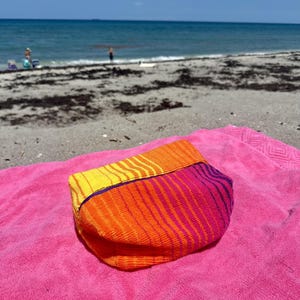 May include: A rectangular pouch made of textured fabric featuring a vibrant gradient pattern that transitions from yellow to orange and pink, accented with thin purple horizontal stripes across the surface and a solid purple trim along the top edge.