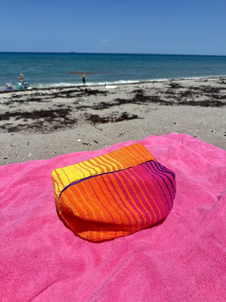 May include: A rectangular pouch made of textured fabric featuring a vibrant gradient pattern that transitions from yellow to orange and pink, accented with thin purple horizontal stripes across the surface and a solid purple trim along the top edge.