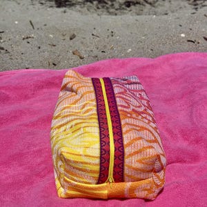 May include: A rectangular zippered pouch featuring a woven fabric with a vibrant orange, yellow, and white abstract pattern. The center zipper is bright yellow, flanked by two decorative bands in deep red and purple with a geometric print.