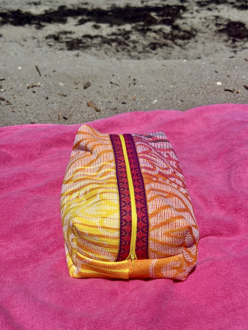 May include: A rectangular zippered pouch featuring a woven fabric with a vibrant orange, yellow, and white abstract pattern. The center zipper is bright yellow, flanked by two decorative bands in deep red and purple with a geometric print.
