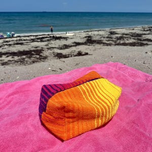 May include: A rectangular zippered pouch made of textured fabric featuring a vibrant color-blocked design. The pouch displays a purple section with thin stripes, followed by a solid orange panel and a yellow section with orange stripes.