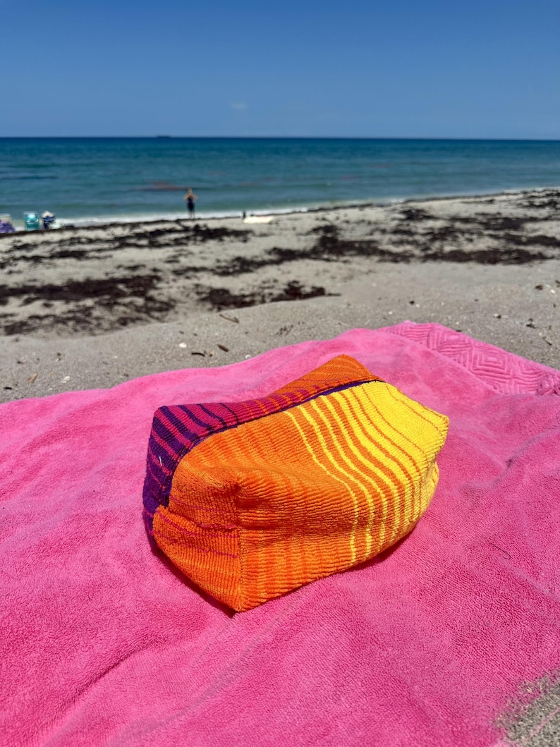 May include: A rectangular zippered pouch made of textured fabric featuring a vibrant color-blocked design. The pouch displays a purple section with thin stripes, followed by a solid orange panel and a yellow section with orange stripes.