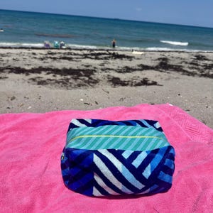 May include: A rectangular zippered pouch featuring a blue, light blue, and white chevron pattern on the sides and a teal and light blue striped top panel with a thin yellow accent line, resting on a bright pink textured fabric surface.