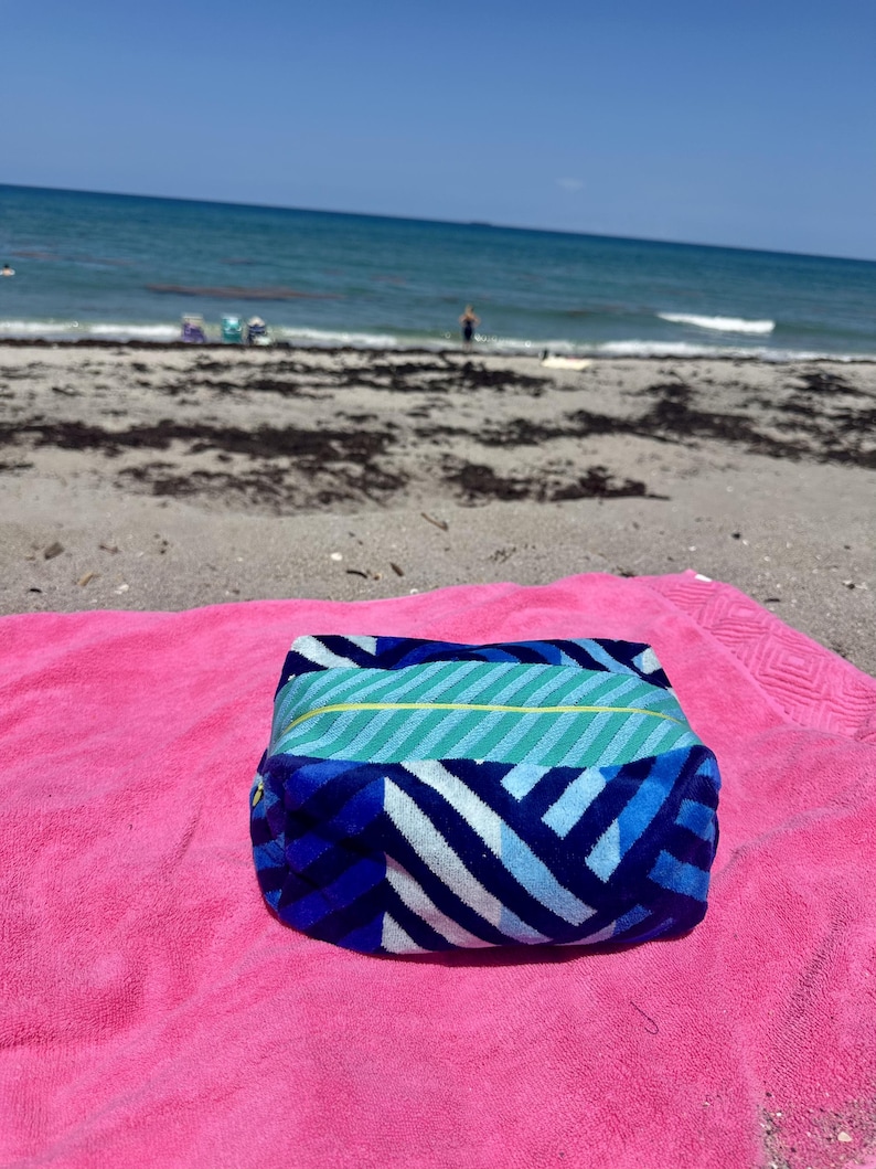 May include: A rectangular zippered pouch featuring a blue, light blue, and white chevron pattern on the sides and a teal and light blue striped top panel with a thin yellow accent line, resting on a bright pink textured fabric surface.