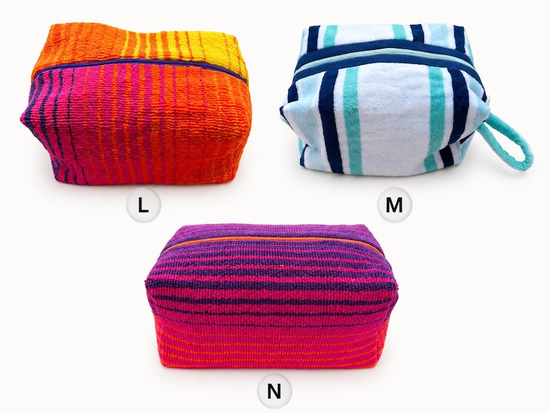 May include: Three rectangular zippered pouches made of textured terry cloth fabric. Pouch L features orange, yellow, and pink stripes. Pouch M has blue, white, and teal stripes with a side handle. Pouch N displays purple, pink, and orange stripes.