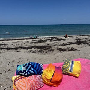 May include: Four rectangular fabric pouches with distinct patterns and colors, including blue, orange, yellow, and pink, resting on a bright pink towel. The pouches feature geometric and wave-like designs in various shades, sitting on a sandy beach.