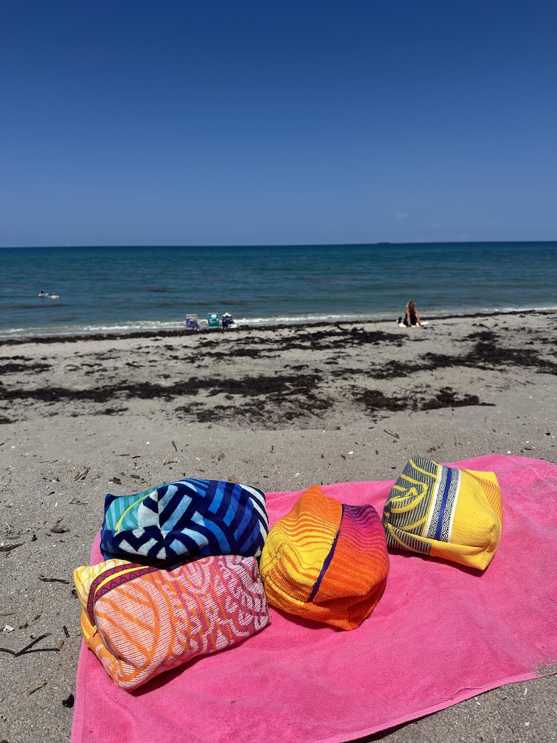 May include: Four rectangular fabric pouches with distinct patterns and colors, including blue, orange, yellow, and pink, resting on a bright pink towel. The pouches feature geometric and wave-like designs in various shades, sitting on a sandy beach.