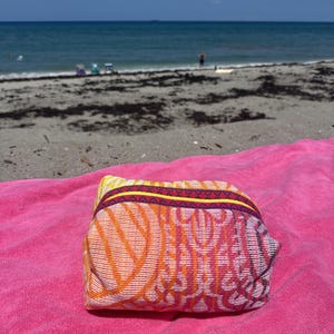 May include: A rectangular zippered pouch featuring a vibrant woven pattern in shades of orange, yellow, and pink. The bag has a distinct yellow and dark purple trim along the top zipper closure and is resting on a bright pink textured towel.