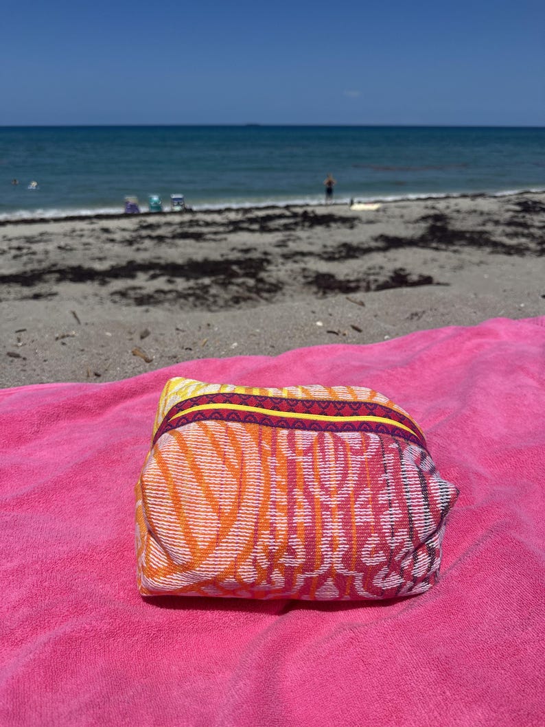 May include: A rectangular zippered pouch featuring a vibrant woven pattern in shades of orange, yellow, and pink. The bag has a distinct yellow and dark purple trim along the top zipper closure and is resting on a bright pink textured towel.