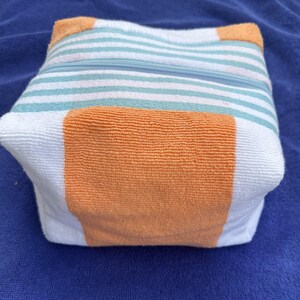 May include: A square-shaped zippered pouch made of terry cloth fabric. The pouch features a pattern of horizontal white and light teal stripes, accented by solid orange panels on the sides and corners. A light blue zipper runs across the top.