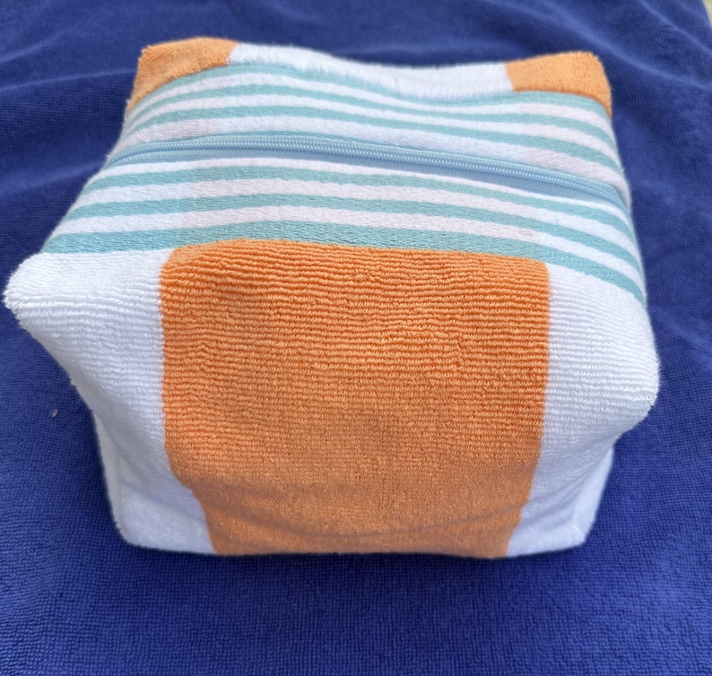 May include: A square-shaped zippered pouch made of terry cloth fabric. The pouch features a pattern of horizontal white and light teal stripes, accented by solid orange panels on the sides and corners. A light blue zipper runs across the top.